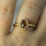 A close-up of a hand wearing a gold ring with an oval faceted yellow gemstone in a textured bezel setting.