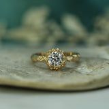 A yellow gold halo ring with a round lab-grown diamond, resting on a textured beige stone surface with a soft-focus teal background.