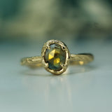 A close-up shot of a gold ring with a textured band and an oval bezel-set yellow sapphire, resting on a light reflective surface with a soft, blurred background.