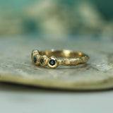 A textured gold ring with small round blue sapphires, resting on a light-colored stone surface with a soft, blurred teal background.