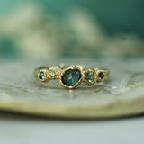 A gold ring with a central rosecut oval sapphire and smaller green and blue sapphires on either side, resting on a textured stone surface with a soft teal background.