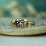 A gold ring with a central rosecut oval australian sapphire and smaller green-teal sapphires on either side, resting on a textured stone surface with a soft teal background.