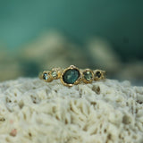A gold ring with a central rosecut oval sapphire and smaller green sapphires on either side, resting on textured white coral with a soft teal background.