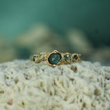 A gold ring with a central rosecut oval sapphire and smaller green sapphires on either side, resting on textured white coral with a soft teal background.