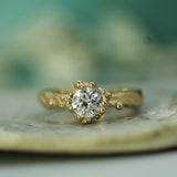 A yellow gold ring with a round-cut lab-grown diamond in a oceany bubbly setting, resting on a textured stone surface with a soft, blurred teal-green background.