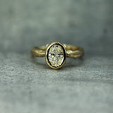 A yellow gold ring with an oval lab-grown diamond set in a bezel setting, resting on a textured gray surface.