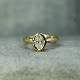 A yellow gold ring with a bezel-set oval lab-grown diamond, displayed on a textured gray surface.