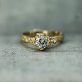 A mermaid treasure style yellow gold ring with a round lab-grown diamond in a oceany bubbly setting, resting on a textured gray surface.