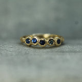 A gold ring featuring five small, round-cut dark blue gemstones set in a textured, organic bezel design, photographed on a neutral gray textured surface.