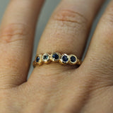 A close-up of a hand wearing a gold band ring with five small round blue sapphires set in a textured bezel-style design.
