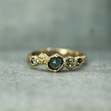 A gold ring with a central rosecut oval australian sapphire and smaller green -teal sapphires on either side, photographed on a textured gray surface.