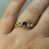 A gold ring with a central dark oval gemstone and smaller round green stones on either side, worn on a person's finger.