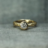 A gold ring with a textured band and a round lab-grown diamond in a bezel setting, resting on a gray textured surface.