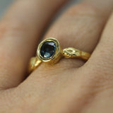 A close-up of a textured yellow gold ring with a oval faceted dark blue sapphire, worn on a person's finger.