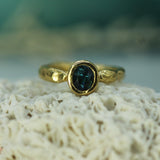 A gold ring with an oval dark blue sapphire, resting on a textured white coral-like surface with a soft, blurred teal background.