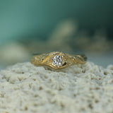 A yellow gold ring with a round lab-grown diamond, displayed on a textured white coral-like surface with a soft teal background.