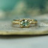 A gold ring with a green oval sapphire in a bubbly setting, resting on a textured stone surface with a soft teal background.