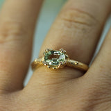 A close-up of a gold ring with a central oval green sapphire and small surrounding accents, worn on a finger.