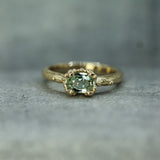 A yellow gold ring featuring an oval-cut light green gemstone in a oceany bubble, resting on a textured gray surface.
