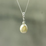 A silver pendant necklace featuring a single pearl with a warm yellow-green iridescence, suspended from a delicate silver chain against a soft, blurred gray-green background.