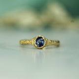 A gold ring with a textured, organic-shaped setting holding a round-cut deep blue sapphire, resting on a light surface with a soft-focus background.