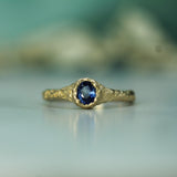A gold ring with a textured, organic-shaped band and an oval blue sapphire in the center, resting on a reflective light surface with a soft, blurred teal background.