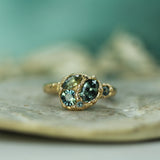 A gold ring featuring blue-green sapphires in a clustered setting, resting on a textured stone surface with a soft teal background.