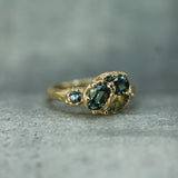 A gold ring featuring blue-green sapphires, with a smaller matching stone on the side, resting on a textured gray surface.