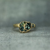 A gold ring featuringblue-green sapphires clustered in the center, photographed on a textured gray surface.