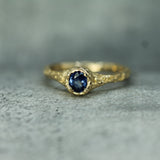 A gold ring with a textured, organic-shaped setting holding an oval deep blue sapphire, photographed on a textured gray surface.