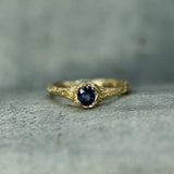 A gold ring with a textured, organic-shaped band and a round dark blue sapphire, resting on a gray stone surface.