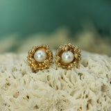 Gold-framed pearl earrings on a textured surface with a blurred natural background