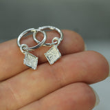 Silver hoop earrings with diamond-shaped pendants held on a hand against a neutral background