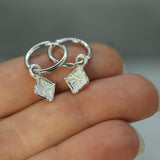 Silver hoop earrings with diamond-shaped pendants held on a hand against a neutral background