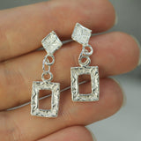 Silver square earrings held between fingers against a neutral background