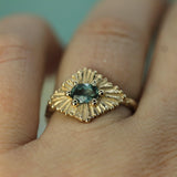 Gold ring with a teal sapphire on on a finger against a light blue background.
