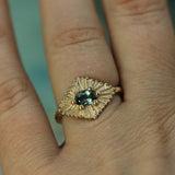 Gold ring with a teal sapphire on a finger against a blurred background