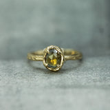A gold ring with a textured, organic-shaped bezel holding a yellow sapphire, resting on a gray stone surface.