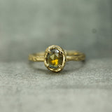 A rustic yellow gold ring with a rosecut oval yellow sapphire, photographed on a textured gray surface.