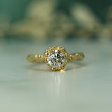 A yellow gold ring with a round-cut lab-grown diamond in a decorative halo setting, resting on a light surface with a soft-focus background.