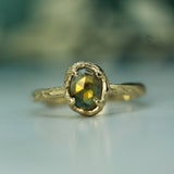 A close-up shot of a gold ring with a textured band and an oval bezel-set yellow sapphire, resting on a light reflective surface with a soft, blurred background.