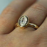 A close-up shot of a gold ring with an oval lab-grown diamond set in a bezel, worn on a person's finger.