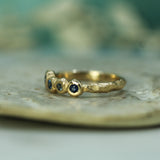 A textured gold ring with small round blue sapphires, resting on a light-colored stone surface with a soft, blurred teal background.