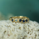 A gold ring with five bezel-set round gemstones, including four dark blue sapphires and one light blue stone, displayed on a textured white coral-like surface with a soft teal background.