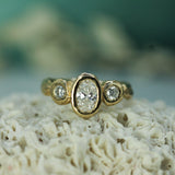 A yellow gold ring with an oval center lab-diamond and two round lab-grown diamonds on either side, displayed on a textured white coral-like surface with a soft blurred teal background.