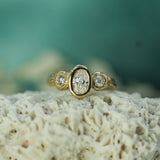 A yellow gold ring with a central oval lab-grown diamond flanked by two round lab-grown diamonds, resting on a textured white surface with a soft teal background.