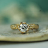 A yellow gold ring with a round-cut lab-grown diamond in a oceany bubbly setting, resting on a textured stone surface with a soft, blurred teal-green background.