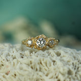 A mermaid treasure three-stone ring in yellow gold with lab-grown diamonds, displayed on a textured white coral-like surface with a soft teal background.