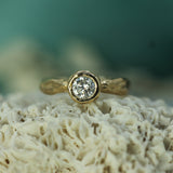 A yellow gold solitaire ring with a round brilliant-cut  lab-grown diamond set in a bezel, resting on textured white coral against a soft teal background.