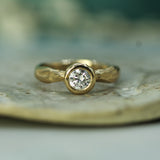 A gold solitaire ring with a round brilliant-cut  lab-grown diamond set in a bezel setting, resting on a textured beige stone surface with a soft, blurred background.